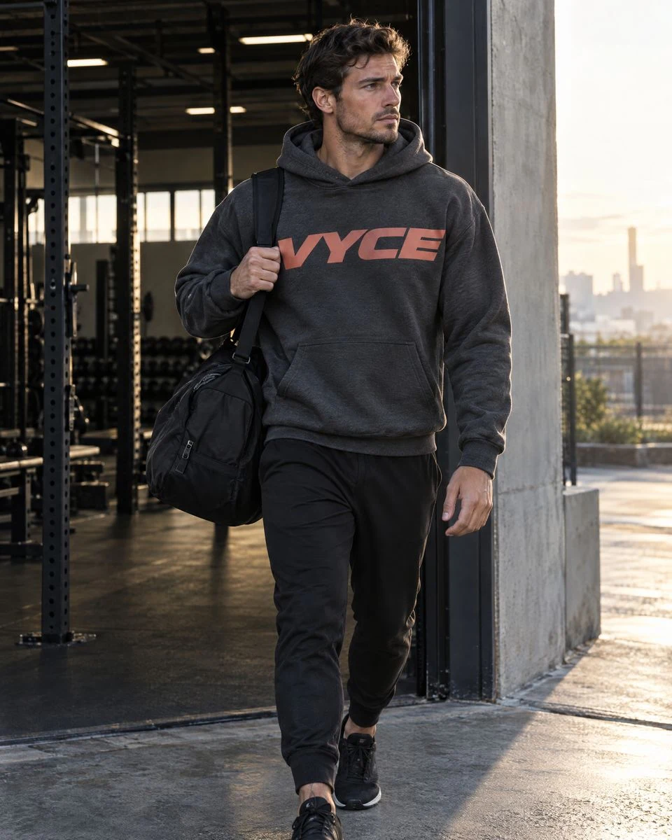 Athlete walking out of an industrial gym at sunrise in a charcoal hoodie with a coral VYCE wordmark across the chest, a black duffel on his shoulder